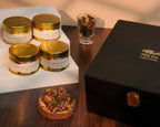 Tea tins with gold lids on a white stand, a glass of tea leaves, and a black box with a brand logo on a wooden surface.