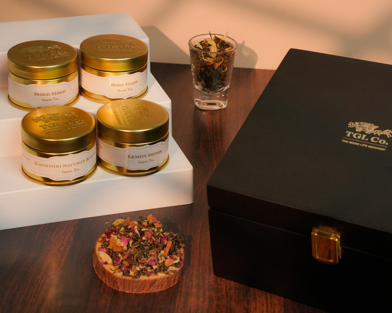 Tea tins with gold lids on a white stand, a glass of tea leaves, and a black box with a brand logo on a wooden surface.