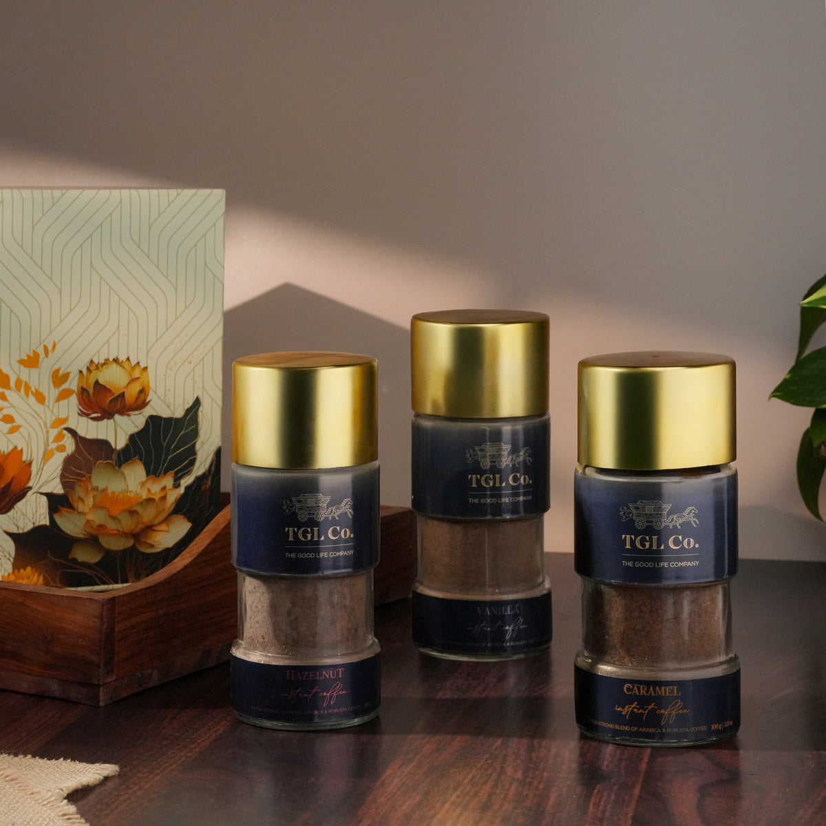 Three spice jars with gold lids on a wooden surface, with a decorative box and plant in the background.